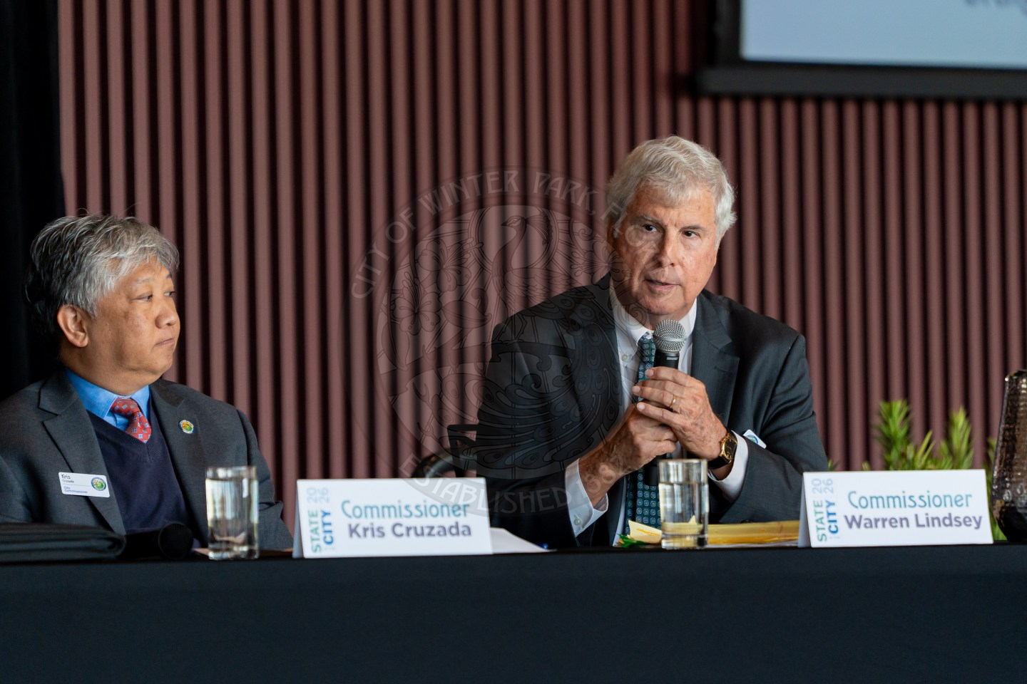 Two commissioners sit at a table during a panel discussion. Commissioner Kris Cruzada listens while Commissioner Warren Lindsey speaks into a microphone. Both have nameplates and water glasses in front of them.