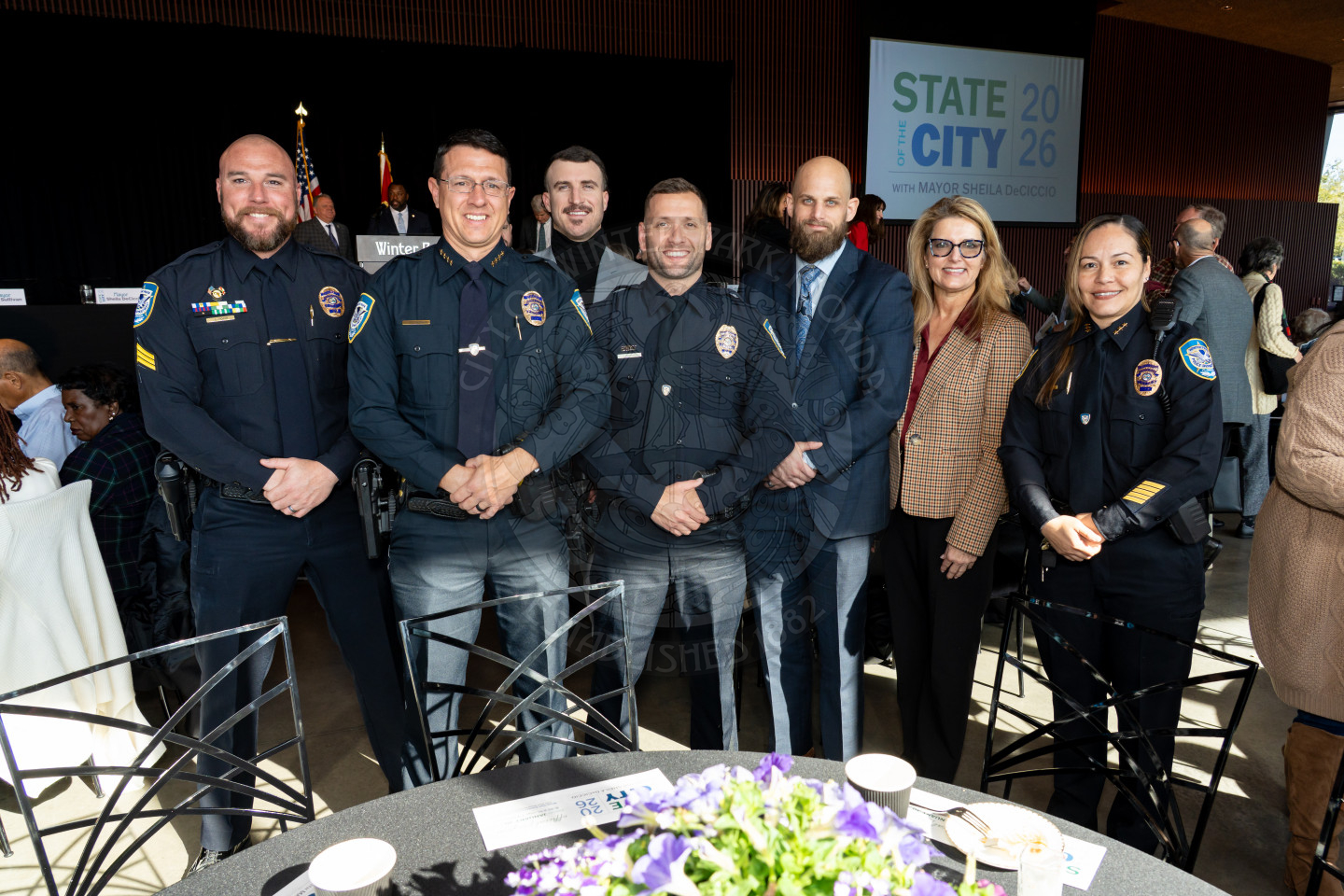 A group of five police officers and two civilians pose and smile together at an indoor event, with a State of the City 2026 sign visible in the background. A table with flowers and papers is in the foreground.