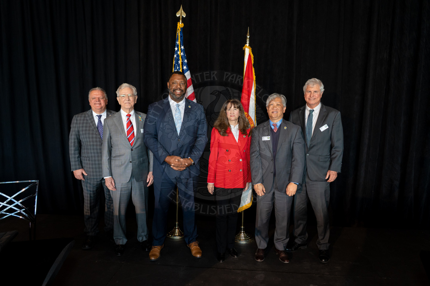 Six people in business attire stand in a row on stage with the U.S. and another flag behind them, posing for a group photo against a black curtain backdrop.