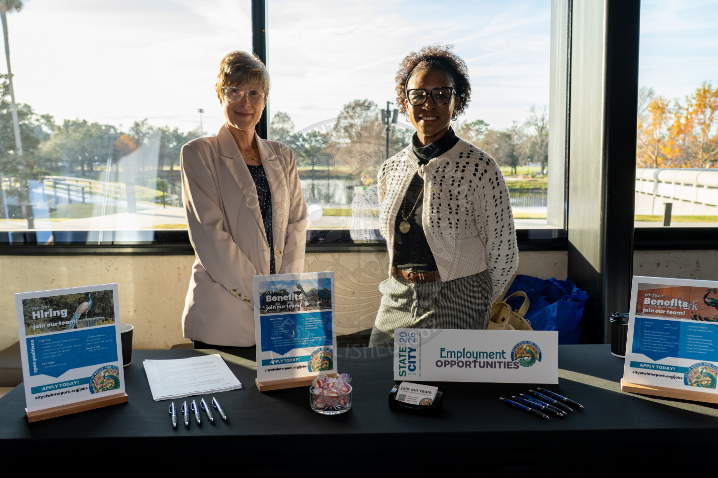 Two women stand behind a table with employment opportunities signs, pens, candy, and informational flyers at a brightly lit indoor event, with large windows showing trees and a pond outside.