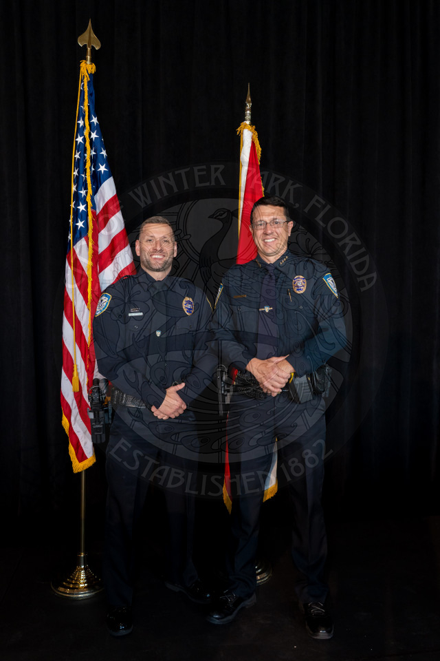 Two uniformed police officers stand smiling in front of U.S. and Florida state flags, with a dark background and a faint city emblem behind them.