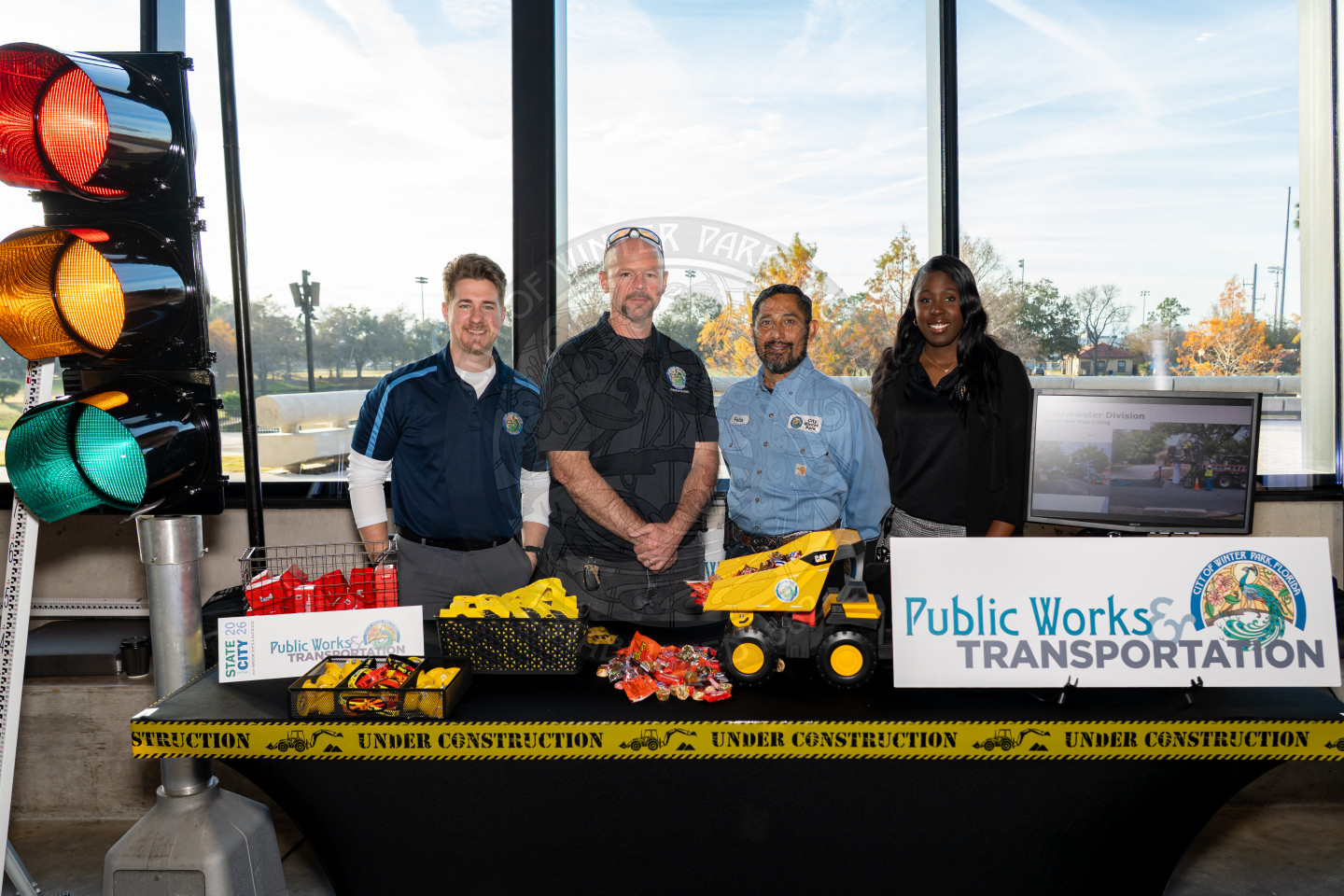 Four people stand behind a table with construction-themed props, Public Works Transportation signs, and giveaways. A large traffic light and Under Construction tape are also displayed.