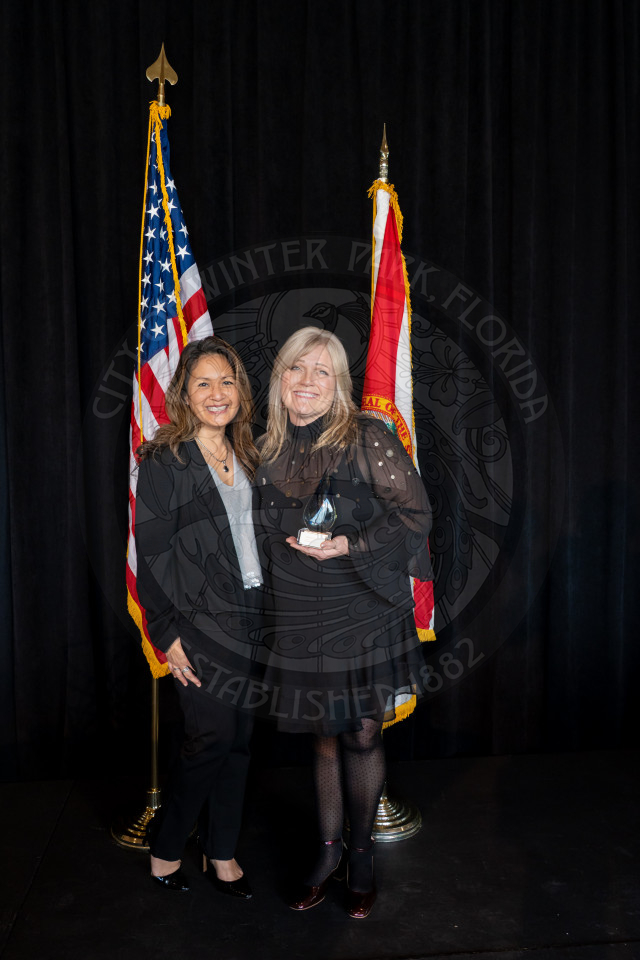 Two women stand smiling in front of the US and Florida flags. One woman is holding a glass award. Both are dressed in formal attire.