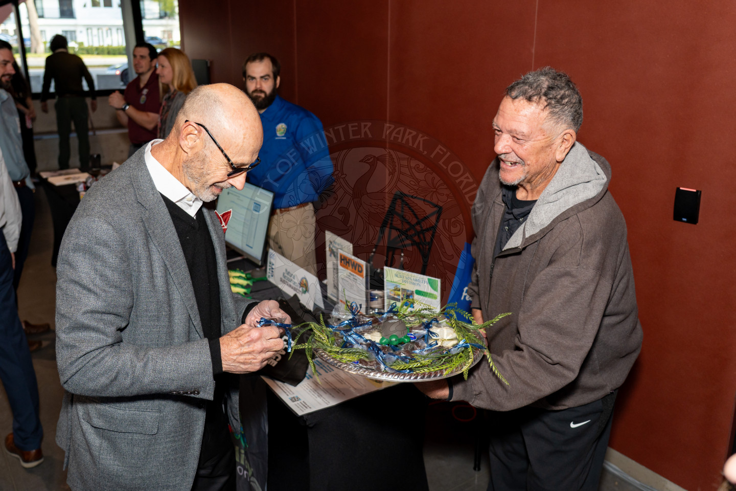 Two men stand at a table with informational displays; one man is handing out chocolate frogs. People and brochures are visible in the background at an indoor event.