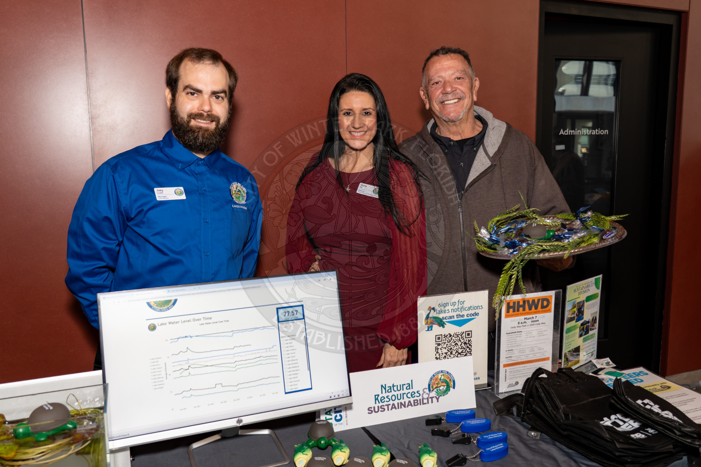 Three people stand behind a display table with environmental sustainability materials, a computer monitor showing data, brochures, and promotional items at an indoor event. They are smiling at the camera.