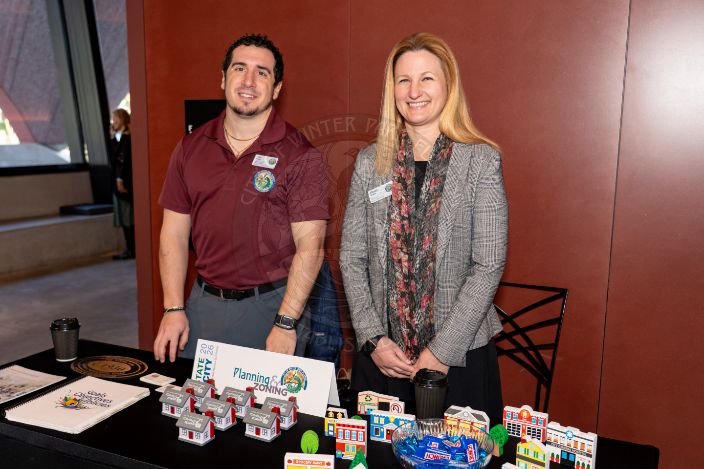 Two people stand behind a table displaying planning and zoning materials, model buildings, and brochures. Both are smiling.