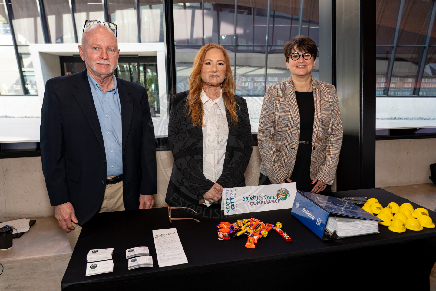 Three people stand behind a table with brochures, candy, business cards, yellow rubber ducks, and folders at an indoor event. Large windows and a modern building can be seen in the background.