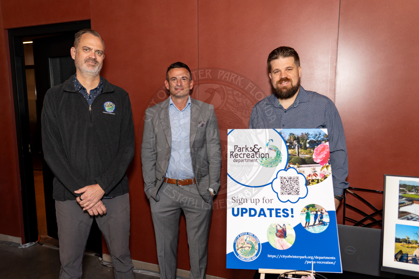 Three men stand indoors near a display board that reads Parks &amp; Recreation Department. Sign up for updates! with a QR code. The board features photos of parks and people outdoors.