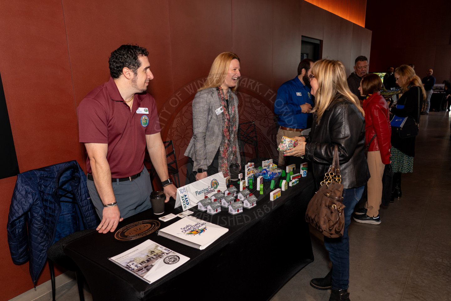 A man and woman stand behind a table with brochures and small toy trucks, talking to a woman in a black jacket at an indoor event. Other people are in the background, engaging with booths.