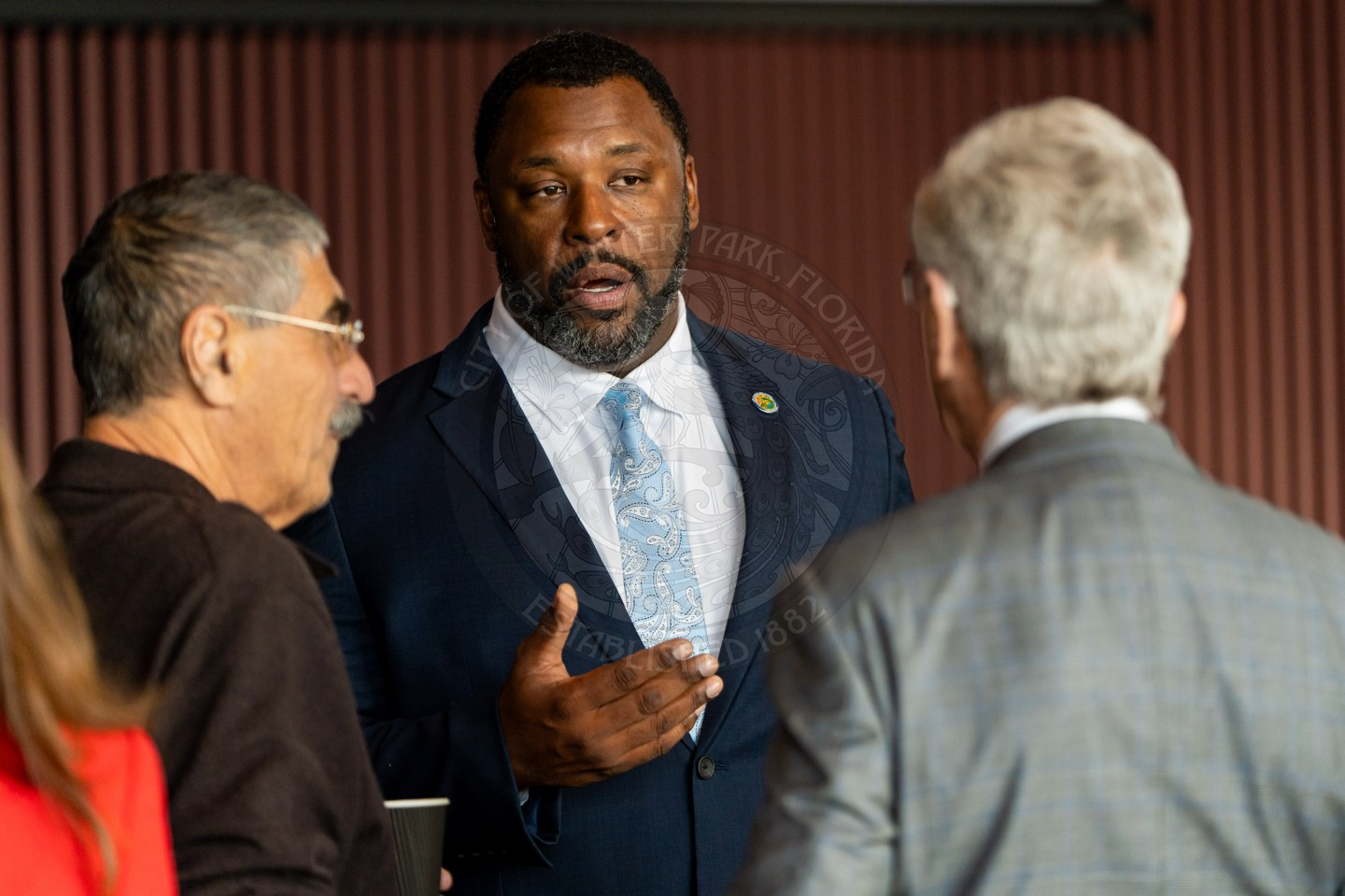 A man in a suit and tie speaks to two other men in business attire, engaging in conversation indoors.