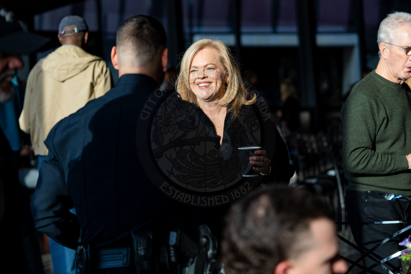 A woman smiles while holding a cup and speaking with a police officer at an indoor event. Other people are gathered around, and sunlight highlights the scene.