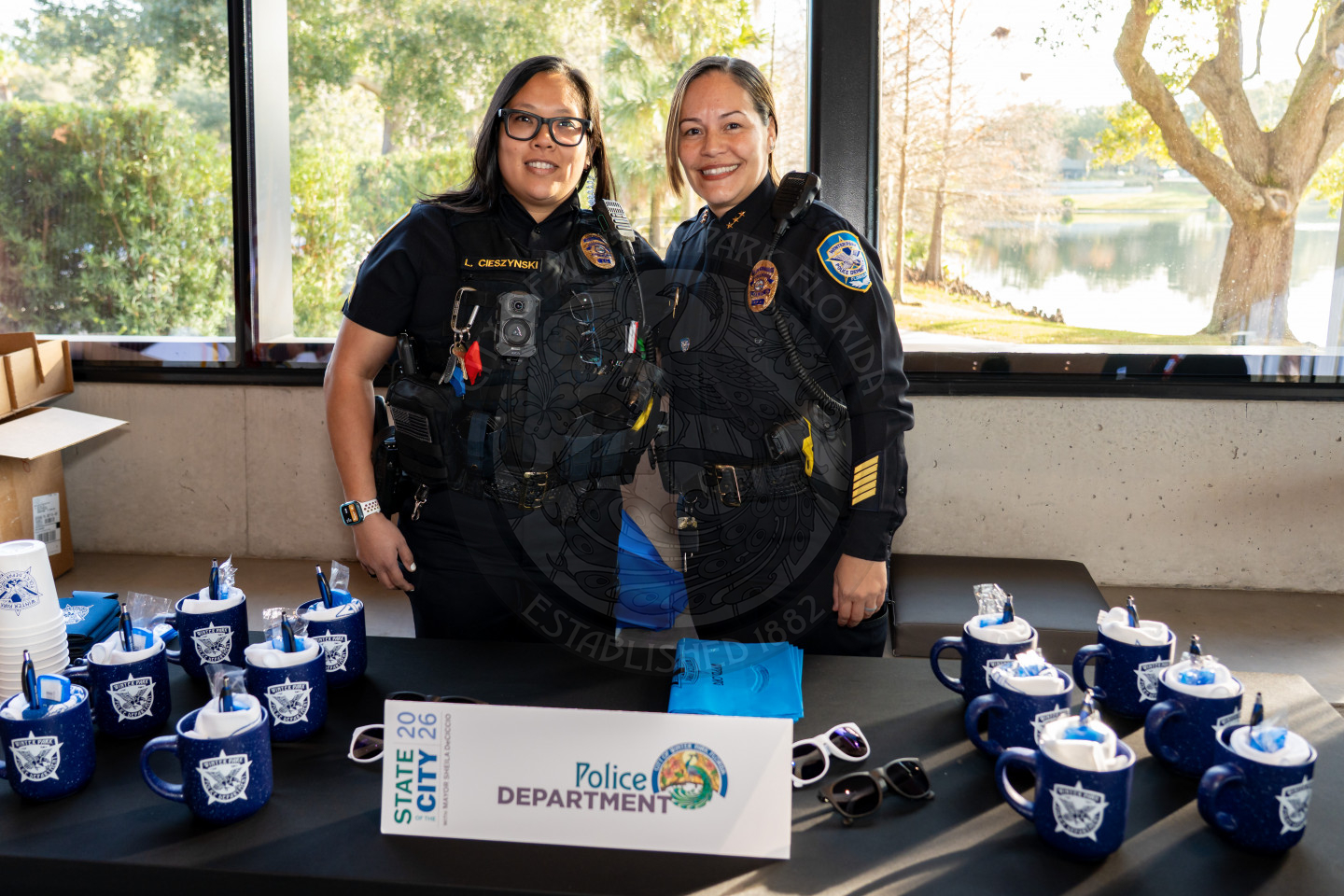 Two smiling police officers stand behind a table displaying mugs, cups, and a “Police Department” sign at an indoor event with large windows behind them and a view of trees and a lake outside.
