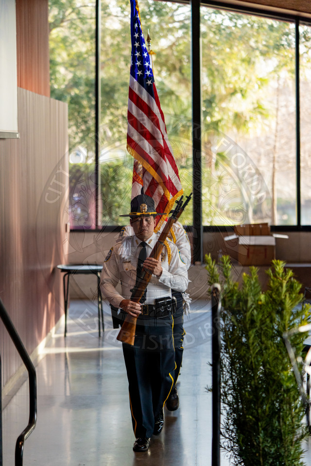Uniformed officers march in a straight line indoors, one holding the American flag, one carrying the Florida State flag and the other carrying a rifle, while sunlight streams through large windows behind them.