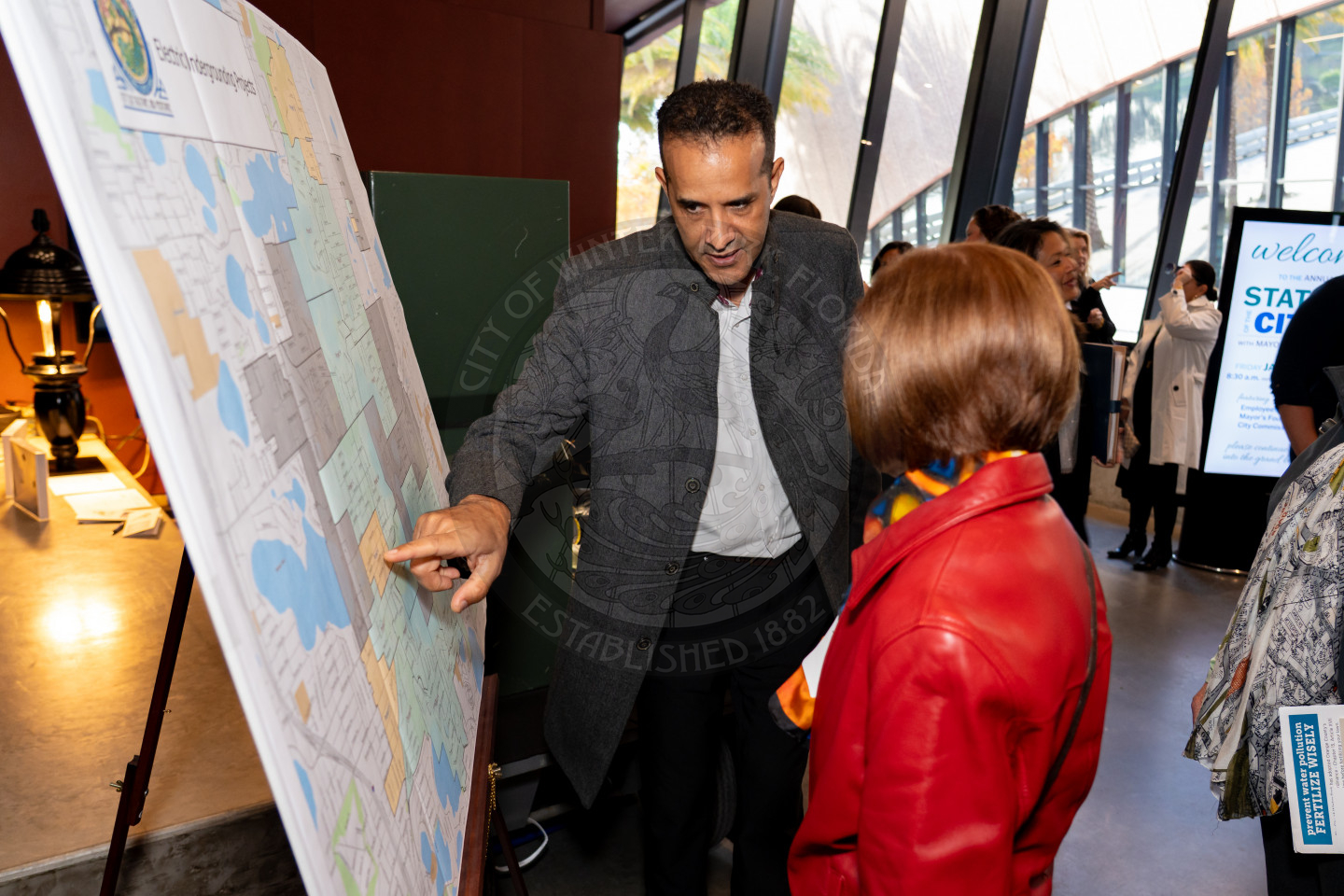 A man points to a large city map on an easel while speaking to a woman in a red jacket at an indoor event, with other people and informational displays visible in the background.