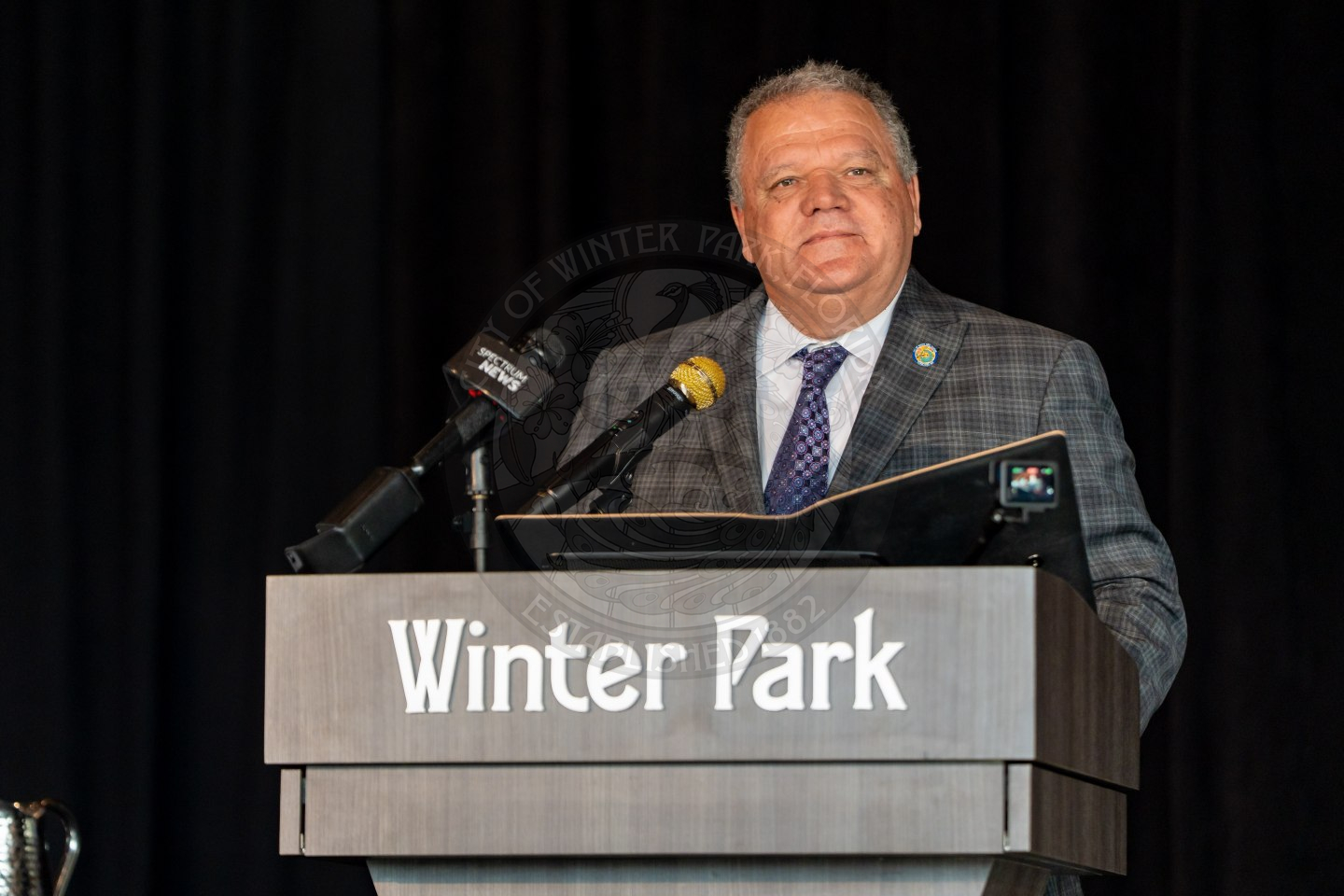 A man in a plaid suit stands at a podium labeled Winter Park, speaking into microphones with a black background behind him.