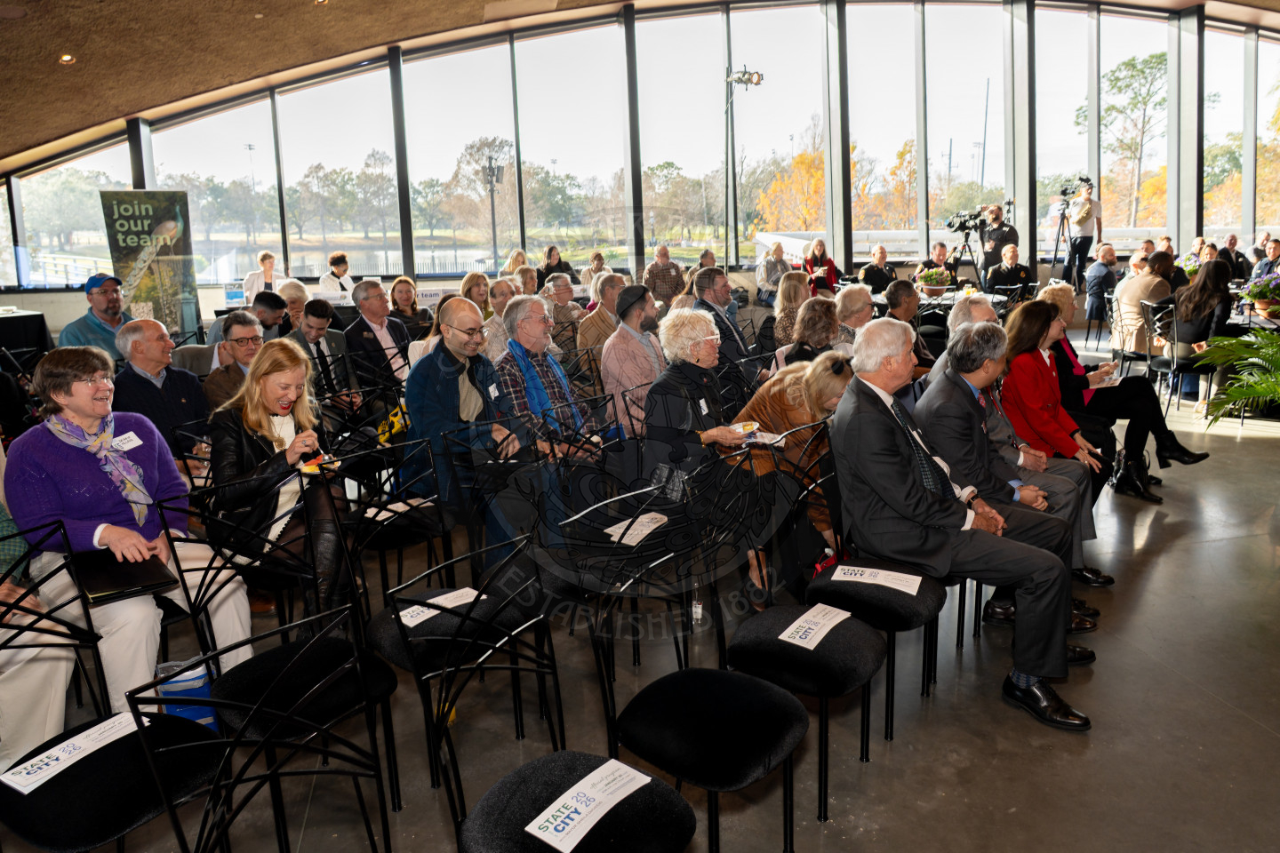 A group of people sit in rows of chairs at an indoor event with large glass windows. Some are talking, while others face forward. A Join our team sign is visible in the background.