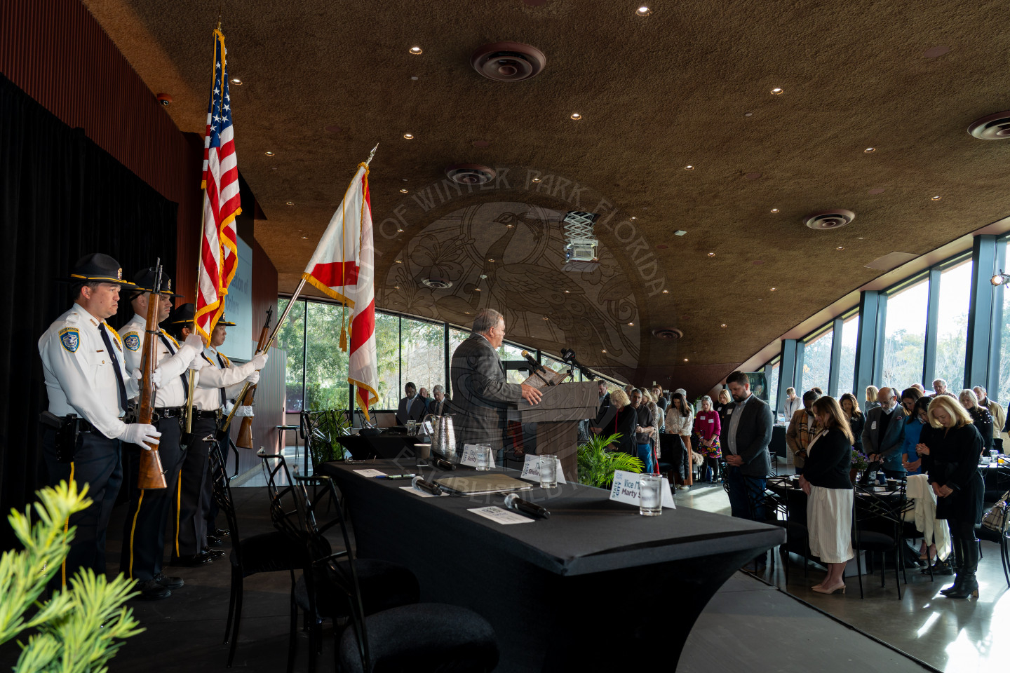 Honor guard holding flags on the stage with a man standing in front of a podium praying. Everyone in the audience has their eyes closed and head down.