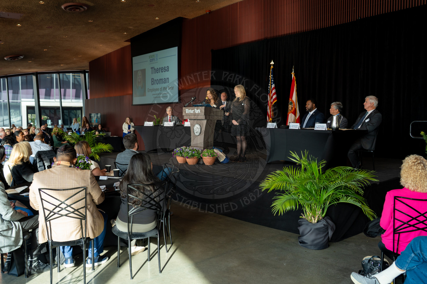 A woman speaks at a podium during an indoor event, with panelists seated at a table onstage, an audience watching, and flags displayed behind the panelists. A presentation slide reads Theresa Bowman on the screen.