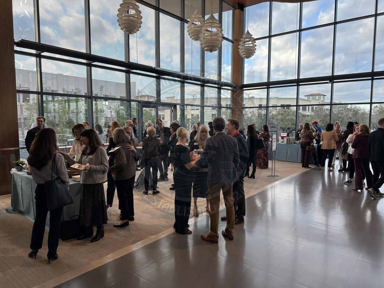 people mingling with each other talking and laughing inside a building with windows from floor to ceiling.