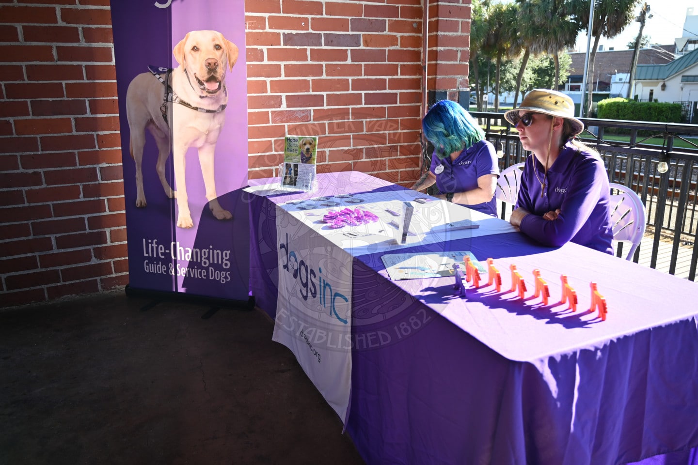 Two people in purple shirts sit behind a Dogs Inc table with a banner featuring a service dog.