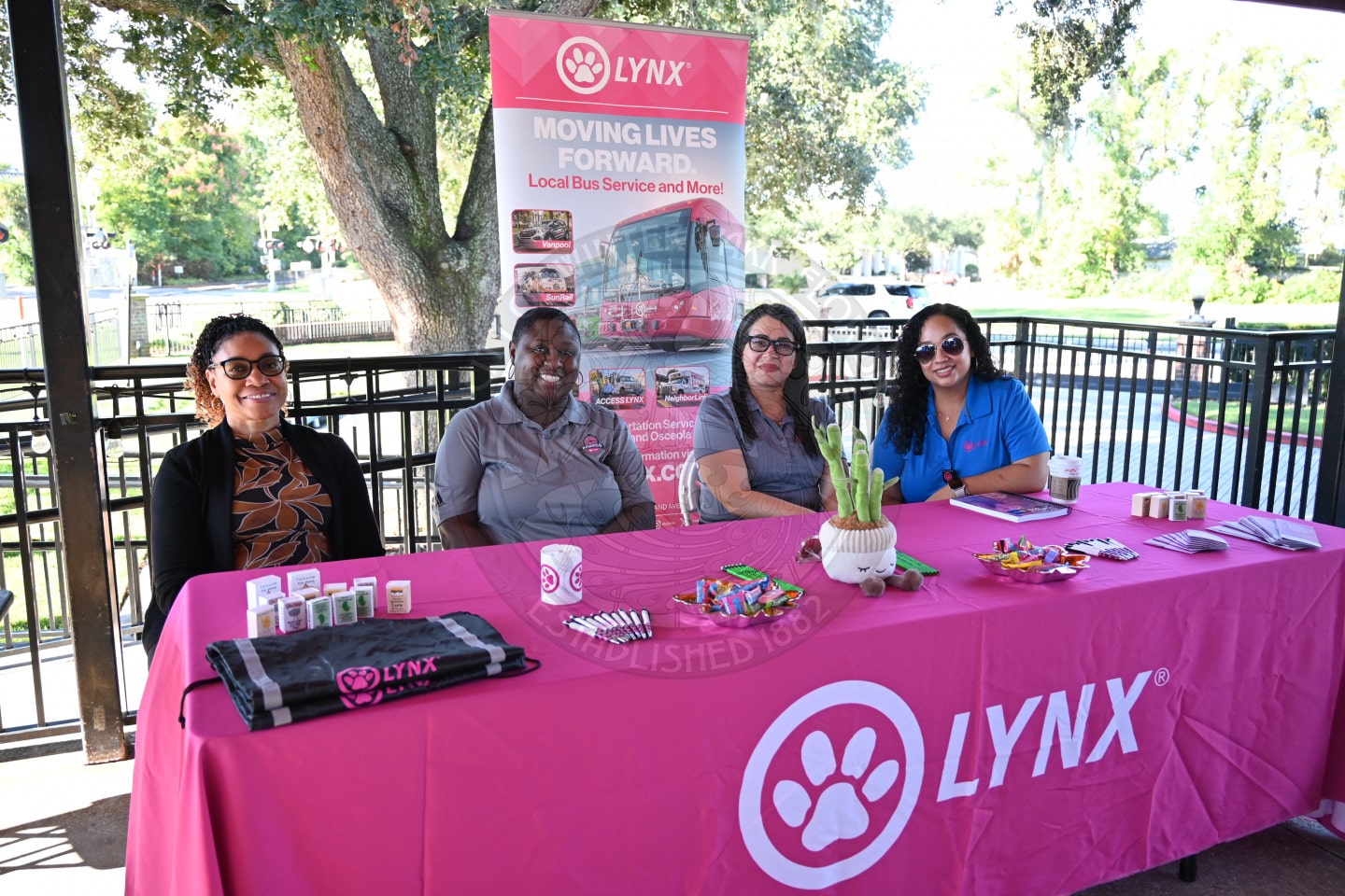Four women sitting behind a pink LYNX branded table at an outdoor outreach event.
