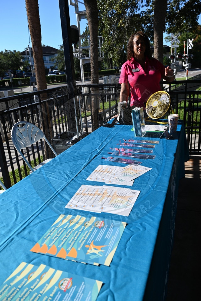 A woman in a pink shirt stands behind a blue-covered table with brochures.