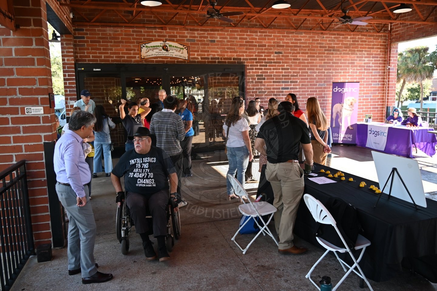 A man in a wheelchair and people gathered at a Dogs Inc informational table under an outdoor brick patio.