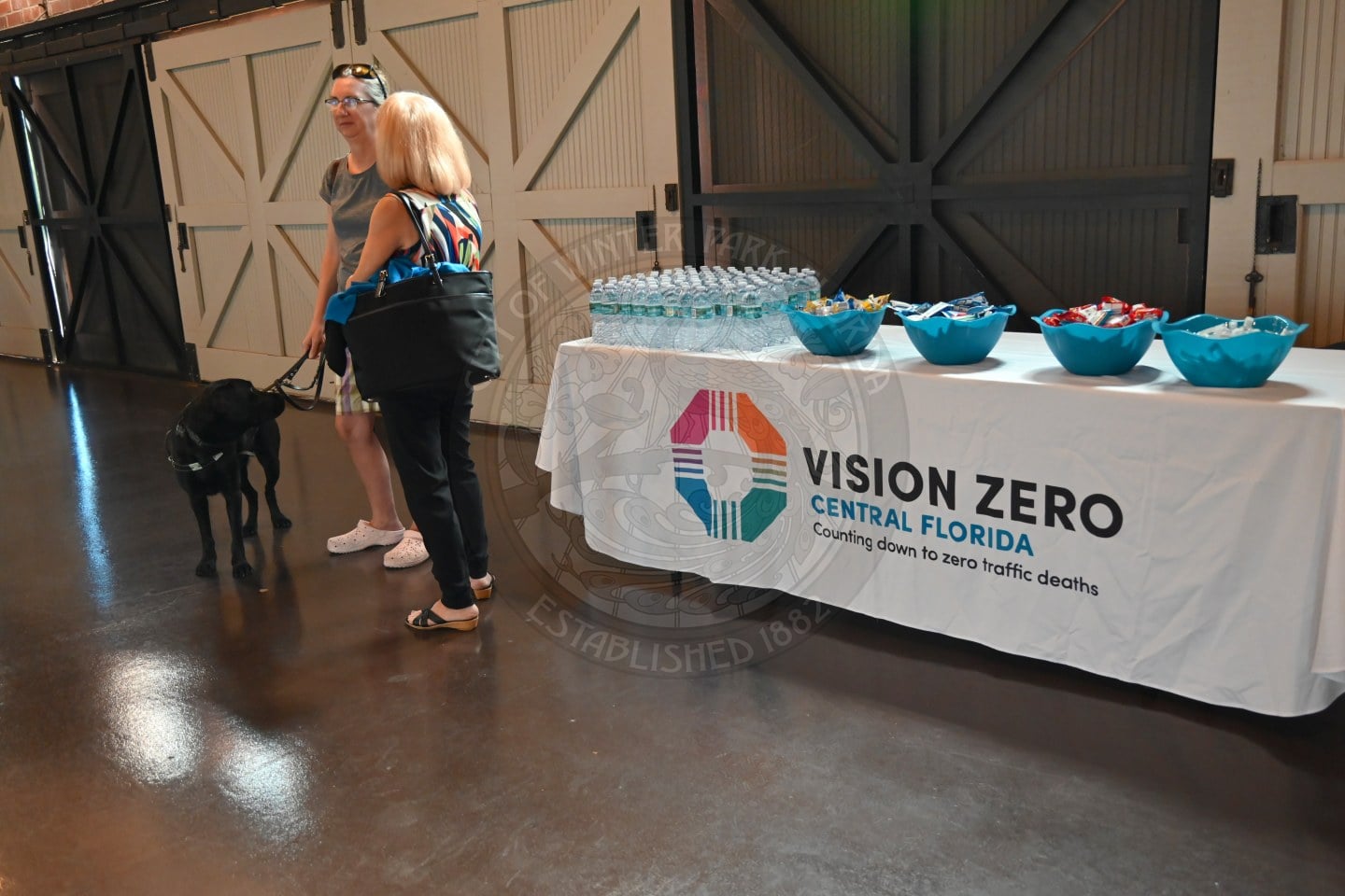 Two people and a black dog stand near a table with a Vision Zero Central Florida banner and bowls of snacks.