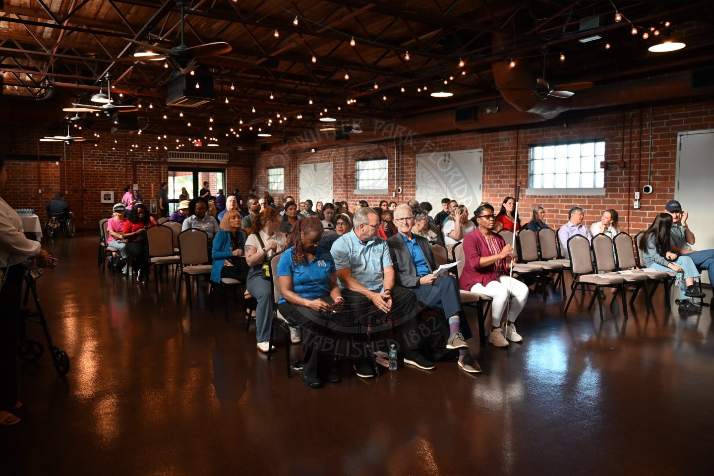 An audience sits in chairs in a large, open room with brick walls and string lights.