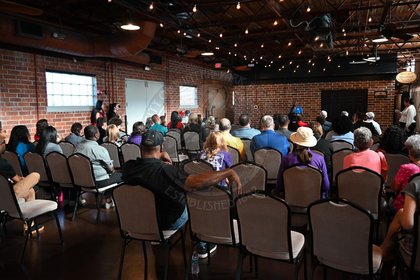 An audience sits in rows of chairs facing a stage inside a brick building with exposed ceiling beams and string lights.