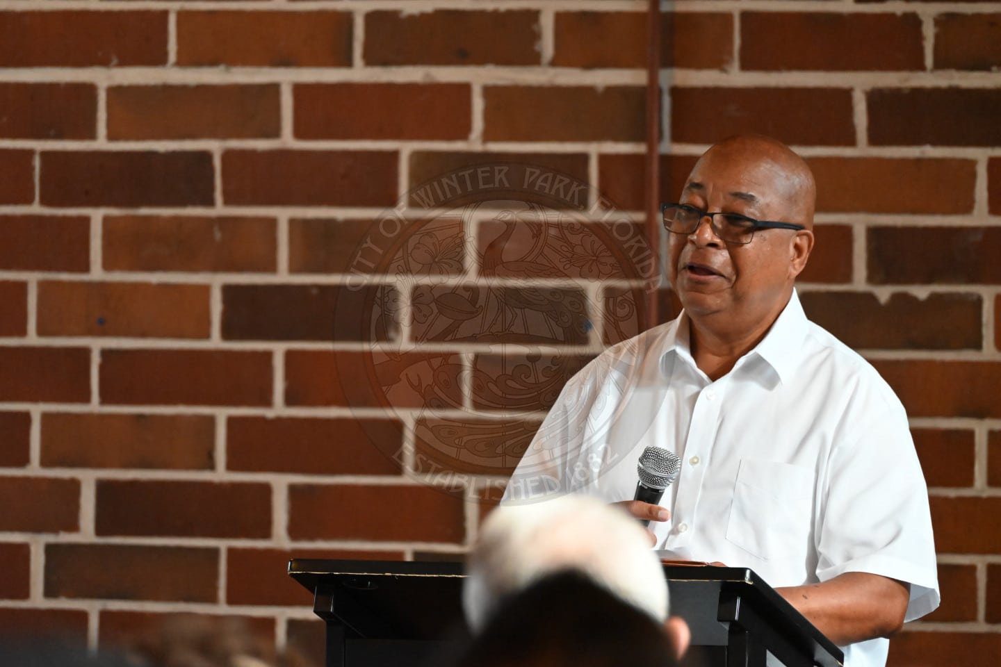 A man in a white collared shirt speaks into a microphone at a podium in front of a brick wall.