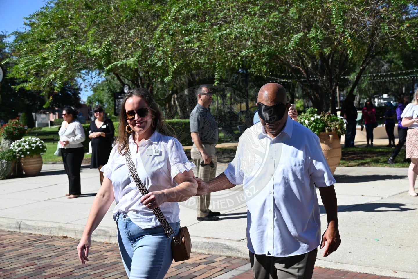 A woman in a white blouse walks outdoors while guiding a man wearing a blindfold.