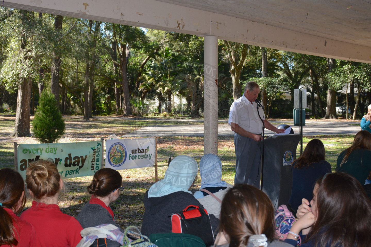 View of Vice Mayor Weldon speaking a the podium with students listening and the Arbor Day banner behind them