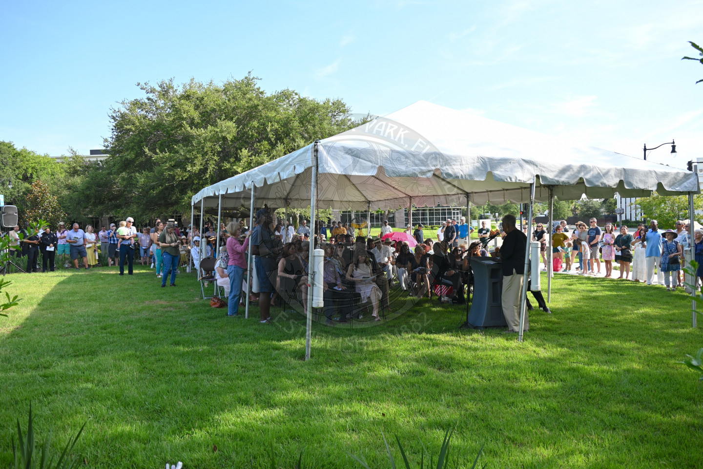 a panaramic view of an event with people sitting and standing around listening to a speaker. 