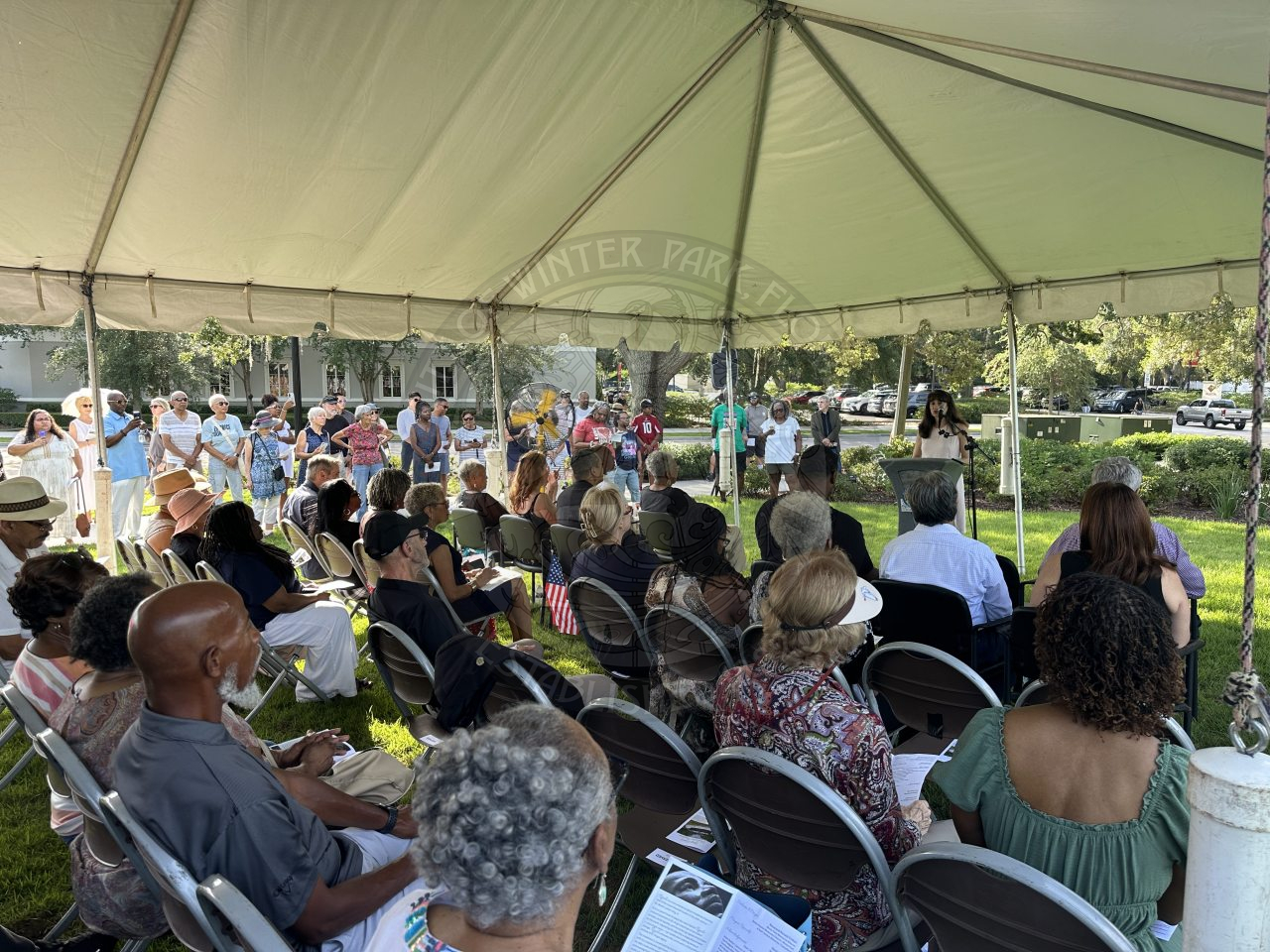 a lady standing behind a podium speaking to a crowd of people sitting in seats watching her.