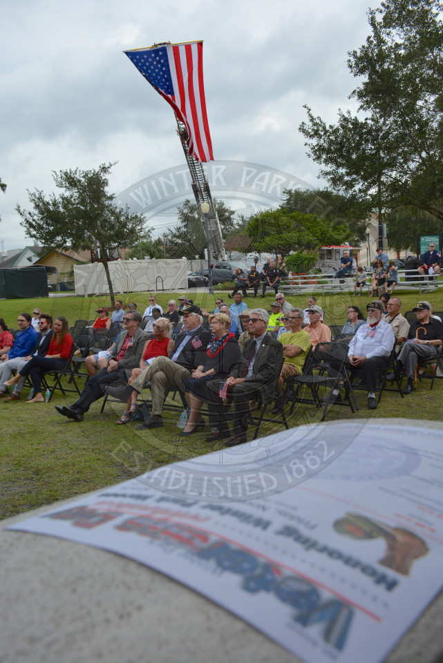 group of people sitting in chairs in shady park watching a veterans day presentation. an american flag is flown in the background