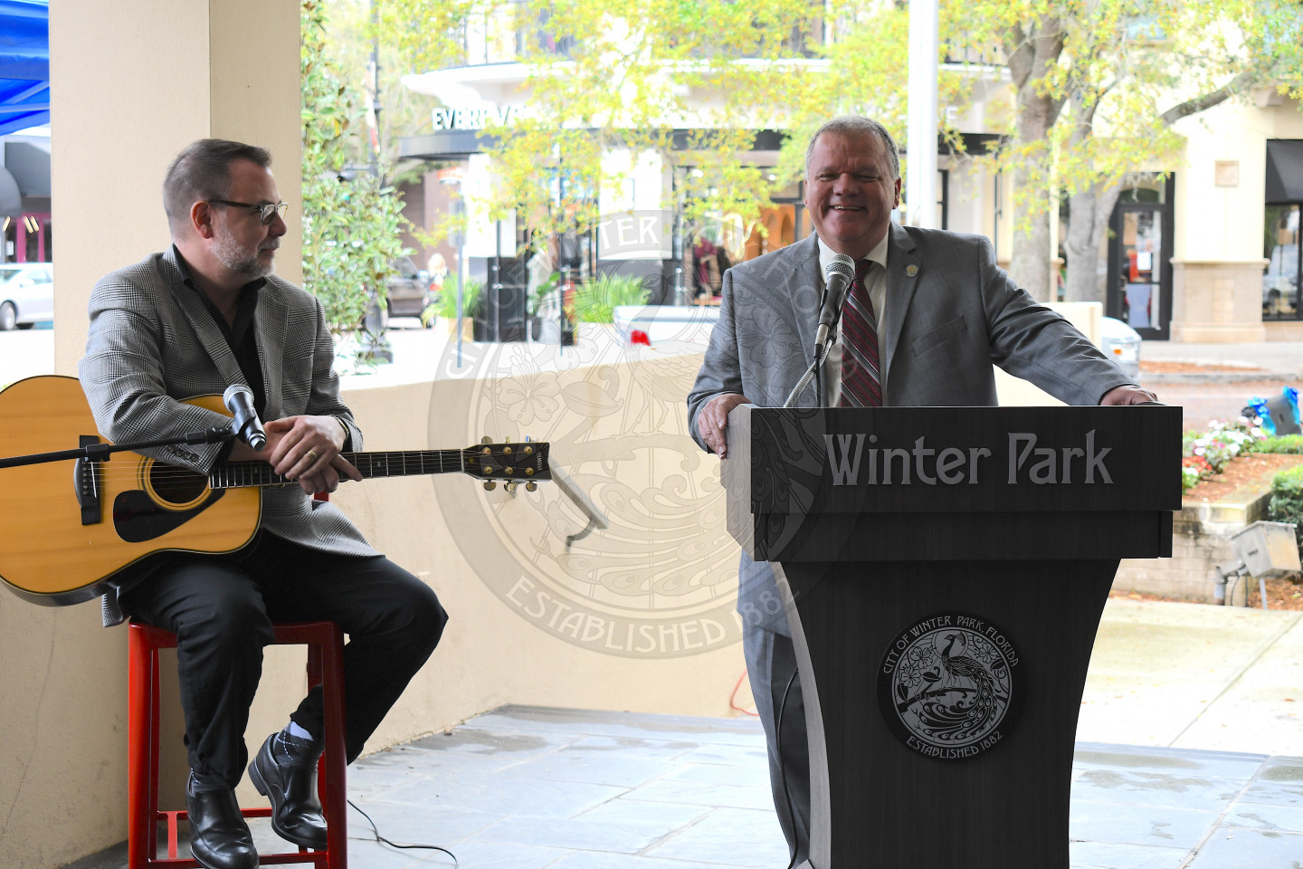 City Manager Randy Knight behind podium talking into microphone with a guitarist sitting in a chair next to him