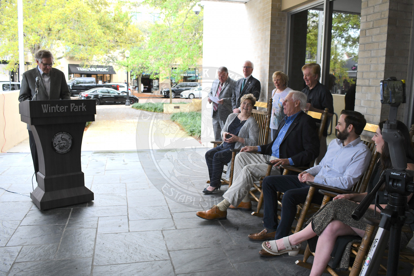 Man standing behind podium talking into microhphone with people sitting in rocking chairs listening to him