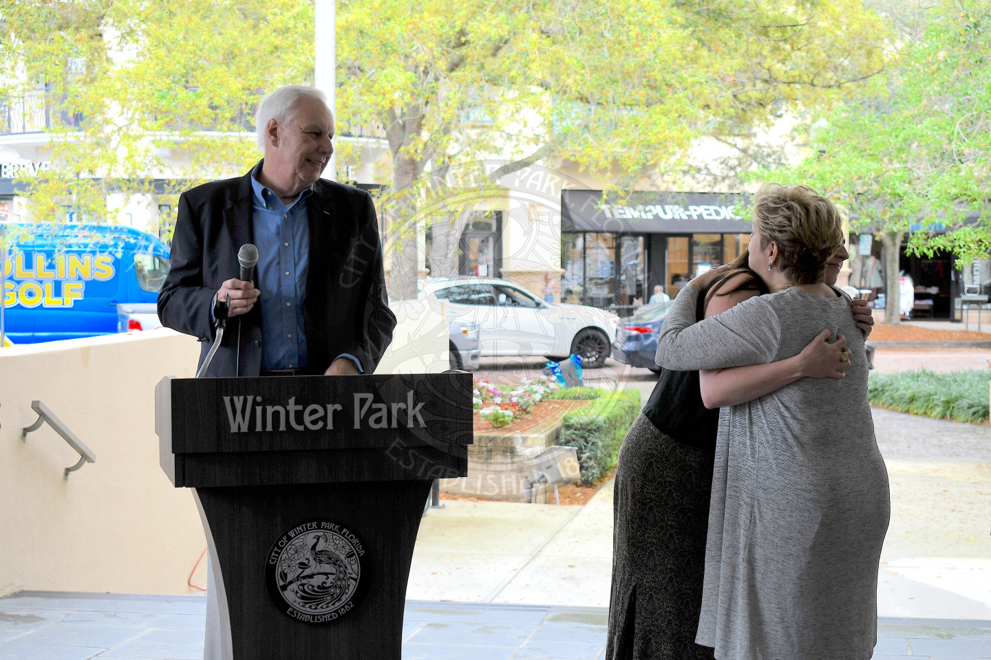 A woman hugging another woman with man standing behind the podium smiling