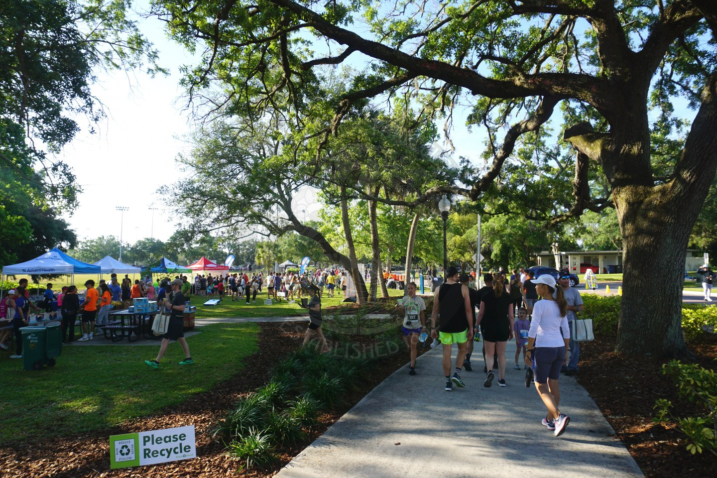 people walking along a path wearing running clothes with 5k racing bibs on them. To the left of the pathway is a grass area with pop up tents for runners to go to after finishing the race.