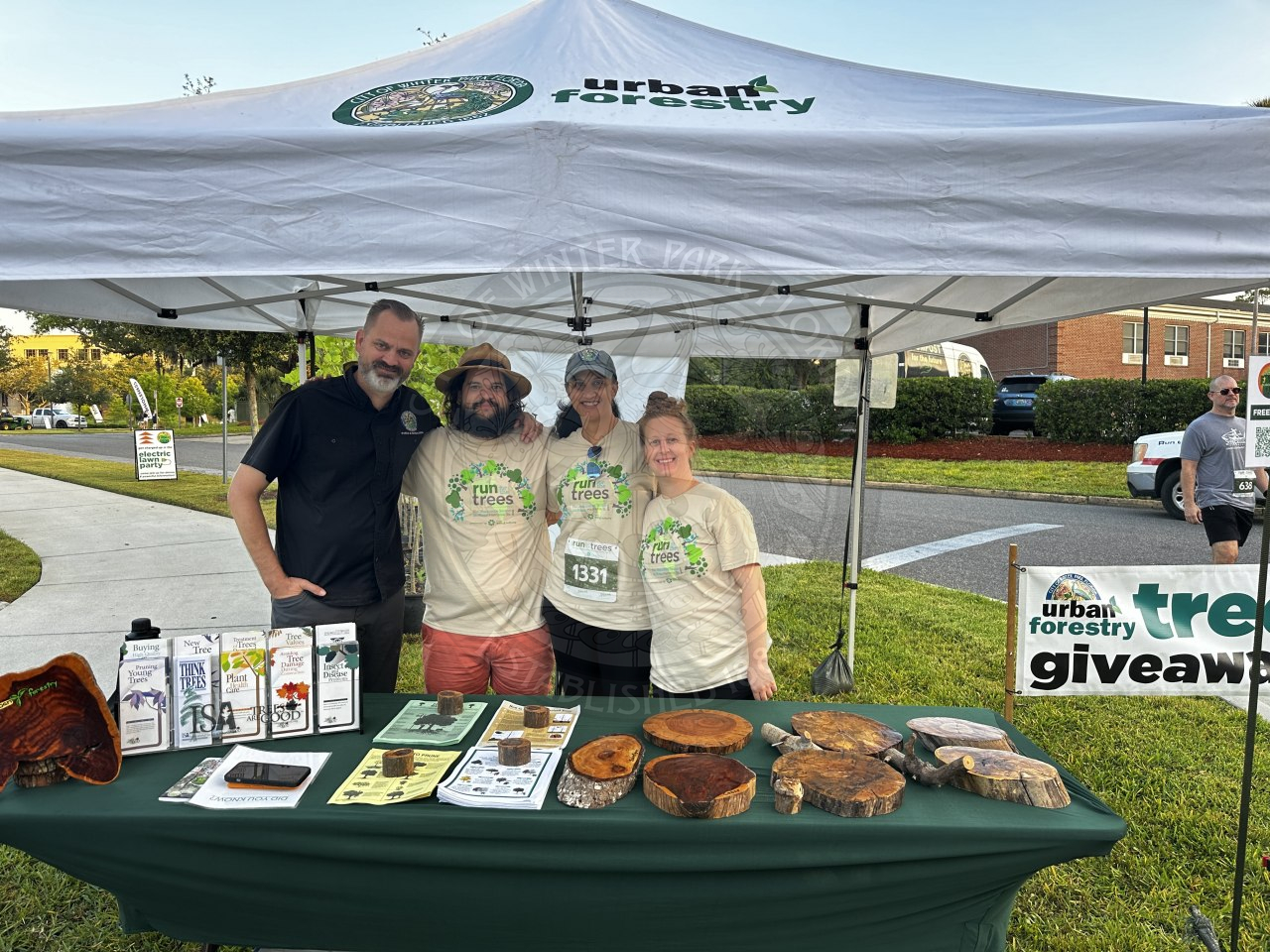 a tent that says urban forestry with the city of winter park city seal. Under the tent is a group of 4 people smiling standing behind a table that has information about plants and trees.