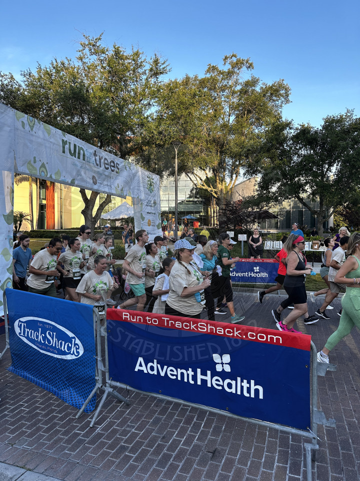 The beginning of a track shack race with everyone running on the road going under a sign that says 'Run for the Trees'. There is barricades on the side with Track Shack and AdventHealth banners.