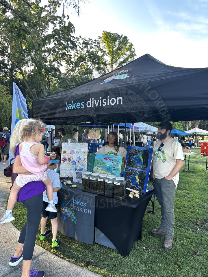 A tent that says lakes division with two city of winter park lakes division staff talking to a lady who is holding a child on her lap. There is a table that has information about winter park lakes on it.