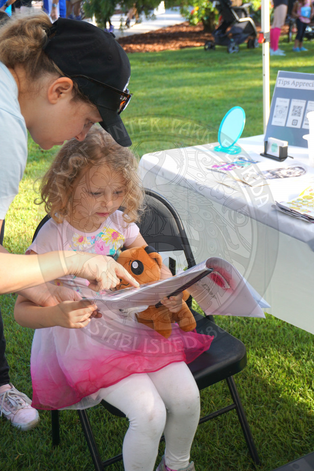 a little girl sitting down holding a book with an adult standing next to her  point at the book