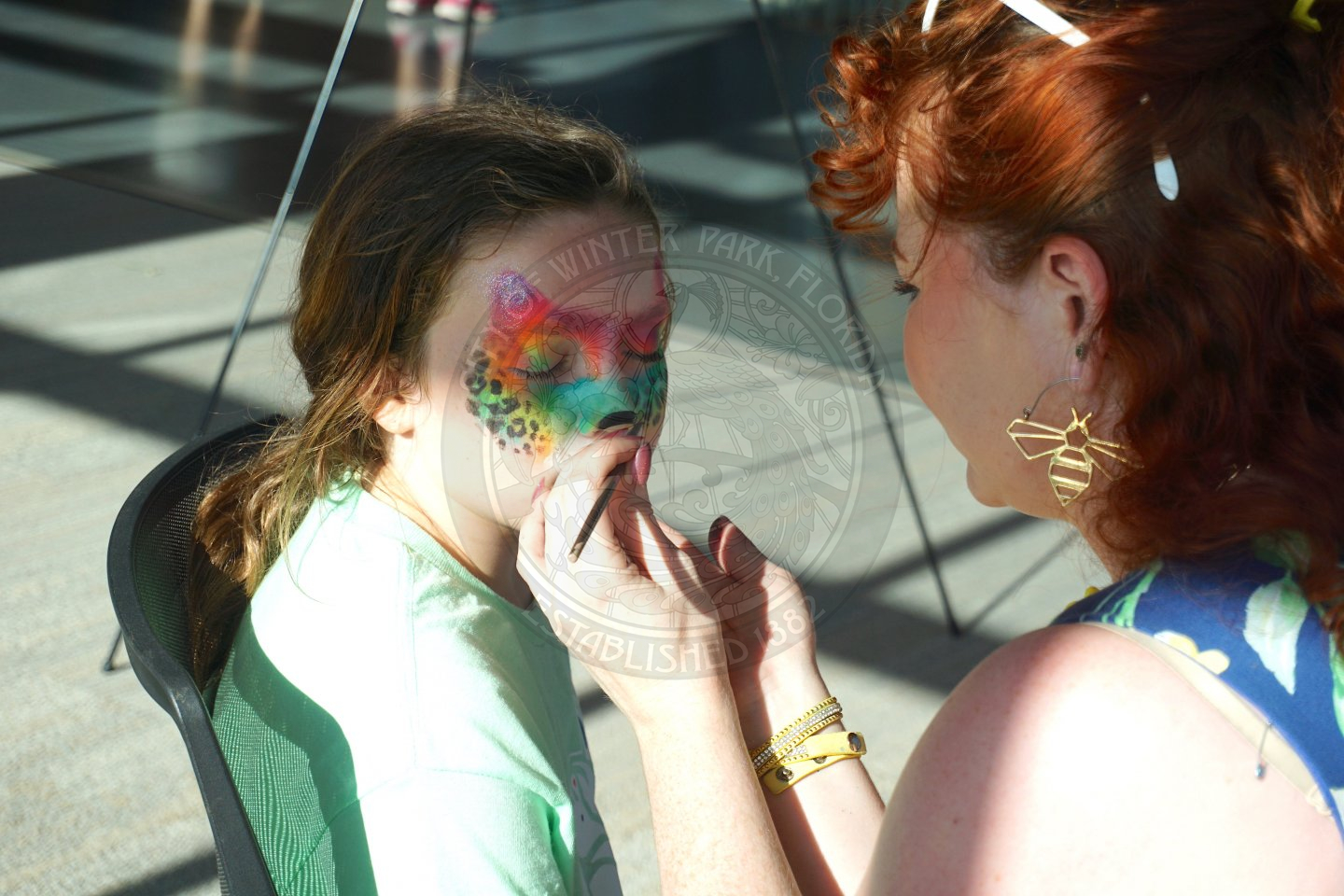 a girl with her face getting painted by a woman with a paint brush in her hand