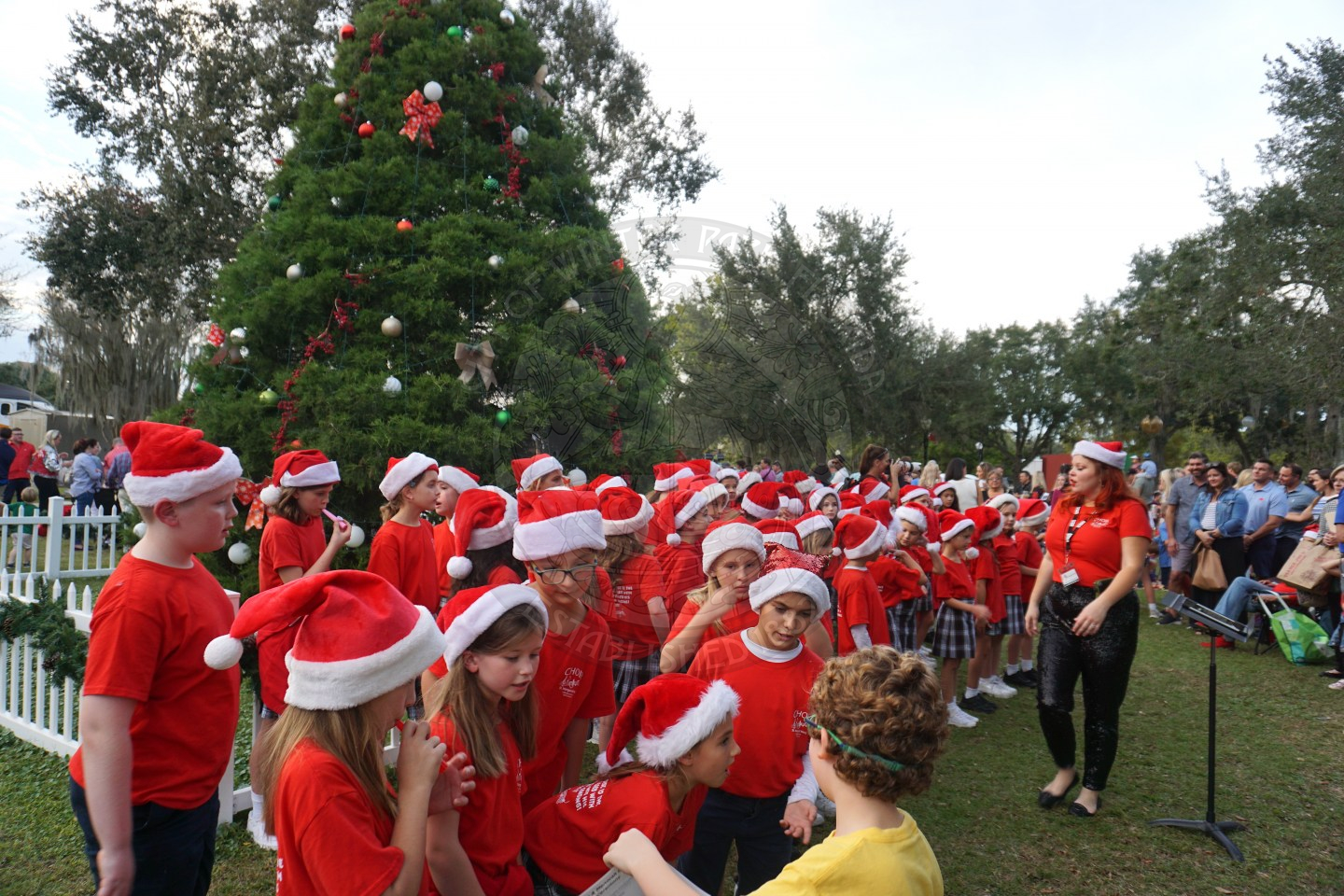 Group of kids all in red shirts and santa hats standing in front of Christmas tree getting ready to sing.