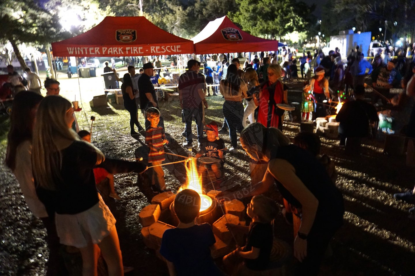 A family standing in a circle surrounding a firepit while holding sticks with marshmallows at one end roasting them in the fire to make smores.