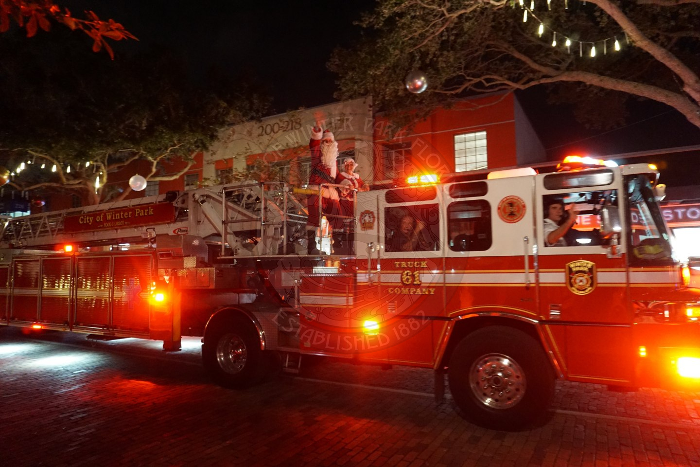 Firetruck driving on Park Avenue with Santa and Mrs. Claus on the top  while santa is waving at all the kids down on the road.