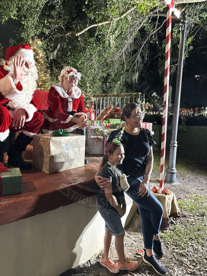 Santa and Mrs. Claus sitting in red chairs on a stage with a bunch of presents surrounding smiling with a mom and daughter standing in front of the stage to take a picture. 