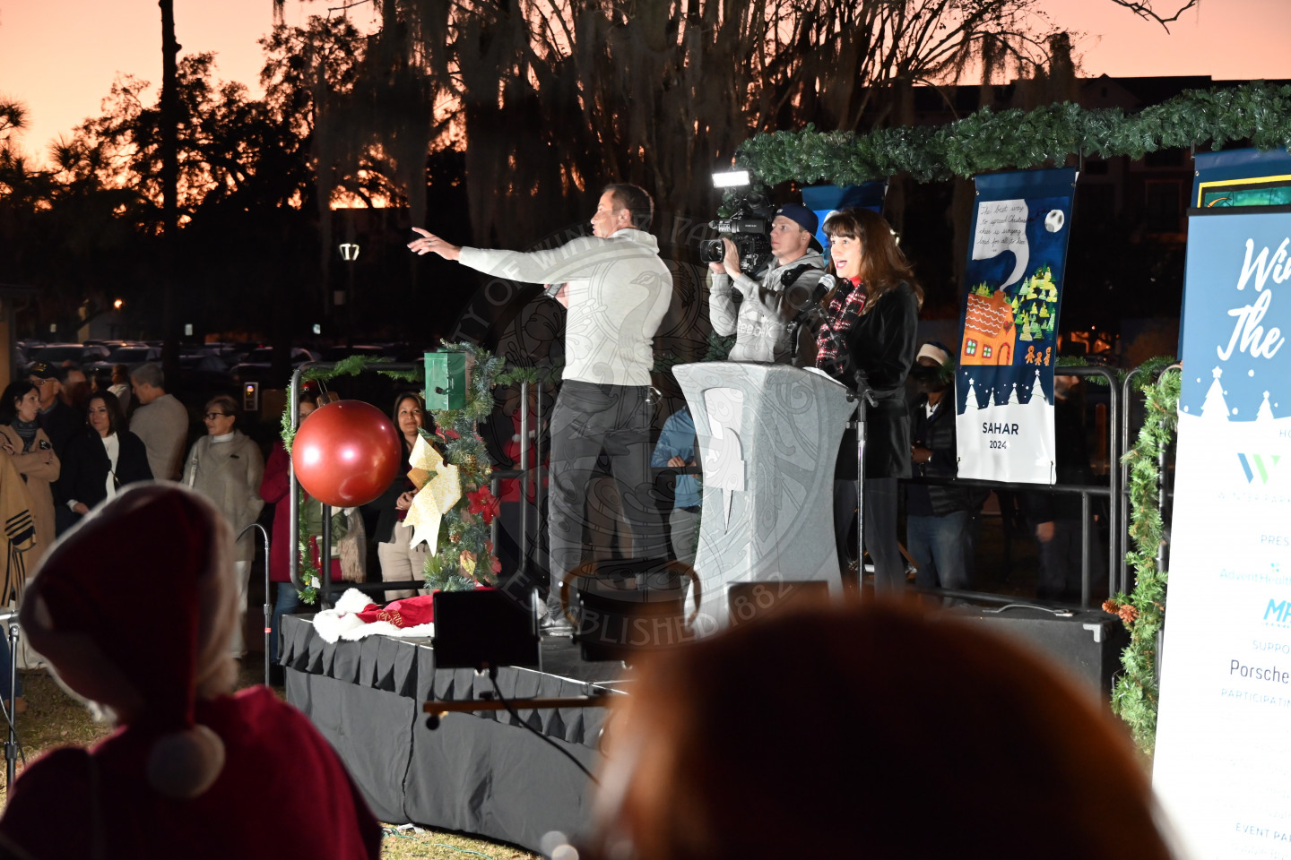 a woman on a stage standing behind a podium speaking to the audience. Next to the woman is a guy talking into a microphone with a man holding a camera filming him.